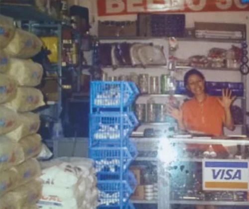 Foto antiga da primeira loja da Trovão: uma mulher acena atrás do balcão, com sacos de cal e cestos de mercadoria