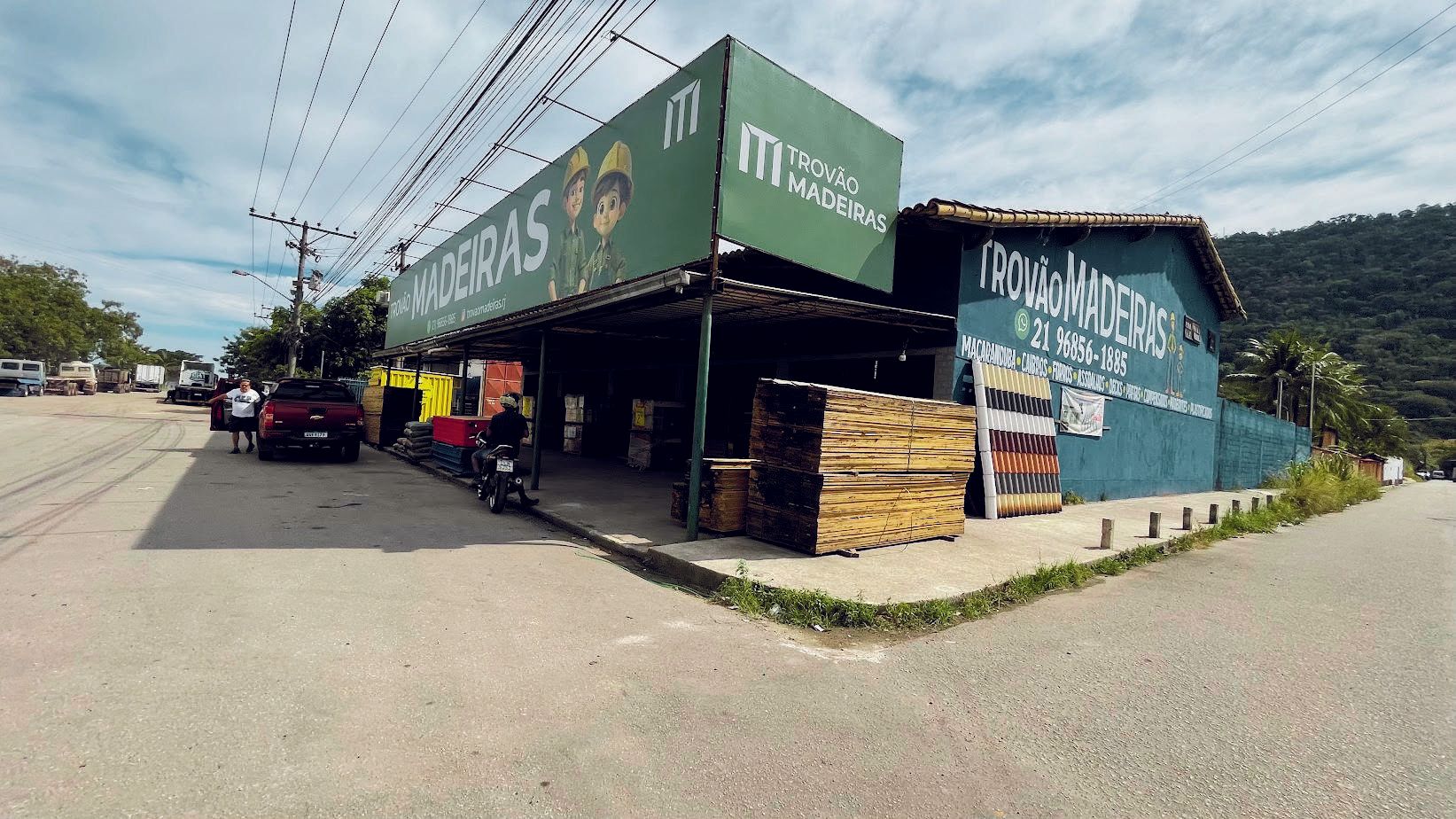 Fachada da Trovão Madeiras com o letreiro verde, madeira empilhada na calçada e o morro ao fundo