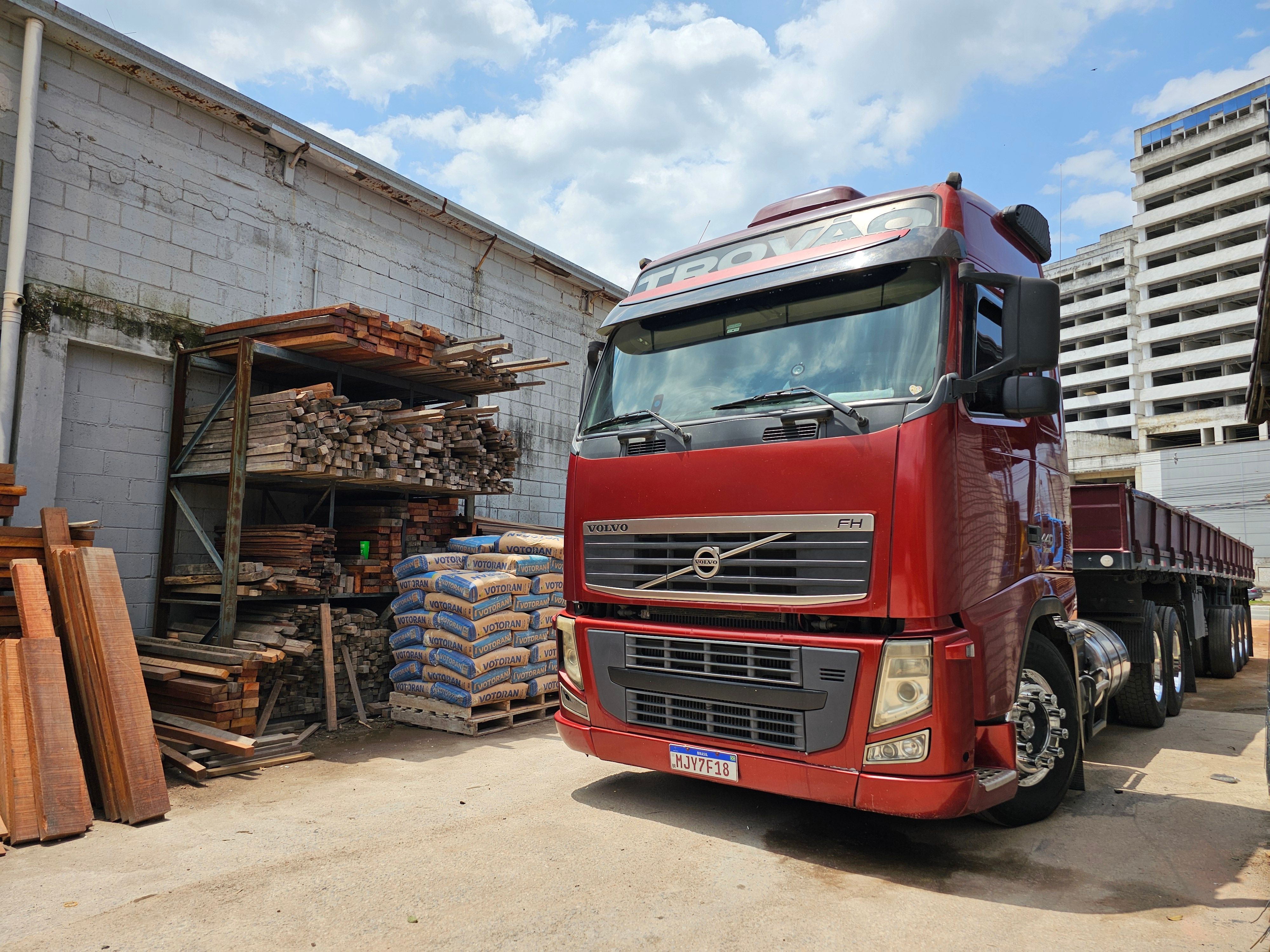 Caminhão Volvo FH da Trovão Madeiras parado no pátio, ao lado das prateleiras de madeira serrada e dos sacos de cimento