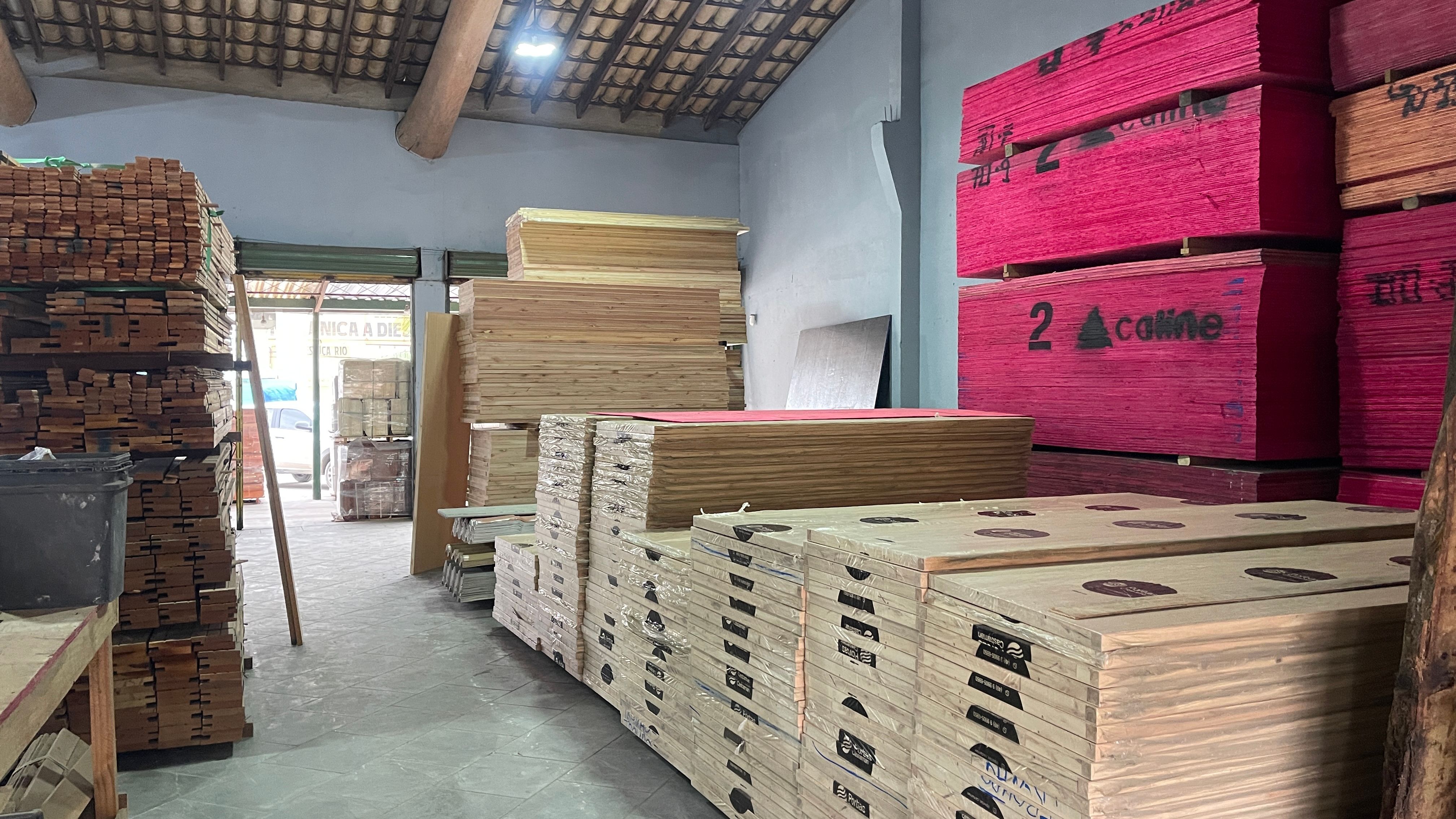 Interior do galpão da Trovão Madeiras: chapas de madeirite plastificado empilhadas à direita e pilhas de tábuas e compensado ao longo do corredor